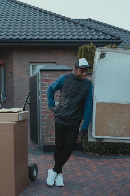 A man with a beard, dressed in a white polo shirt, jeans, and a black cap, is standing inside the open rear of a white moving van, which is parked outdoors on a paved driveway or street. He is holding