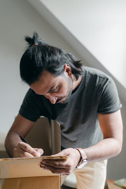 A man with dark hair tied in a small ponytail is inside a home, leaning over a cardboard box, and carefully sealing or taping its edge during packing for a house move. He is wearing a dark grey t-shirt and a wristwatch, working near a window with natural light illuminating the scene. The room has a plain wall and a window frame visible in the background, suggesting an indoor environment typical of residential packing and moving preparations. The presence of packing materials and the focused action supports the context of home relocation, as the man arranges boxes and prepares items for transport. This image exemplifies the packing process involved in house removals, highlighting careful handling and organization, which are essential steps in furniture transport and moving logistics managed by services such as those provided by Man with Van Dulwich.