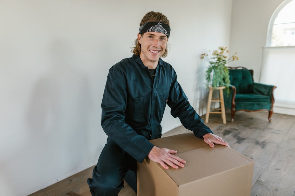 A man with shoulder-length blond hair and a cheerful expression, wearing a dark blue work uniform and a black headband, is seen inside a well-lit room during a house move. He is in the process of packing or preparing a large cardboard box, which he is holding steady with both hands. The room features wooden flooring, a green upholstered armchair with wooden legs, and a small wooden side table with a potted plant and white flowers on top. Natural light enters through a large arched window in the background, illuminating the space. This scene captures the loading and packing stage of home relocation, illustrating furniture and cardboard packing materials commonly used by services like Man with Van Dulwich for removals and moving logistics.