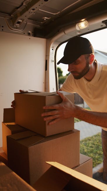 A man with dark hair tied in a small ponytail is inside a home, leaning over a cardboard box, and carefully sealing or taping its edge during packing for a house move. He is wearing a dark grey t-shirt and a wristwatch, working near a window with natural light illuminating the scene. The room has a plain wall and a window frame visible in the background, suggesting an indoor environment typical of residential packing and moving preparations. The presence of packing materials and the focused action supports the context of home relocation, as the man arranges boxes and prepares items for transport. This image exemplifies the packing process involved in house removals, highlighting careful handling and organization, which are essential steps in furniture transport and moving logistics managed by services such as those provided by Man with Van Dulwich.