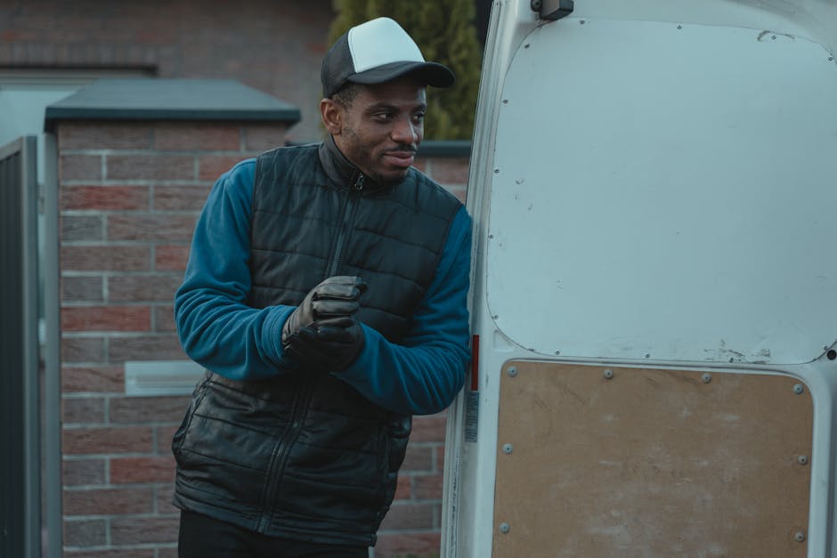 A man dressed in a dark quilted vest over a blue long-sleeved top, wearing a black and grey cap, stands outside a white van with an open rear door. He is holding a small object in his gloved hands and