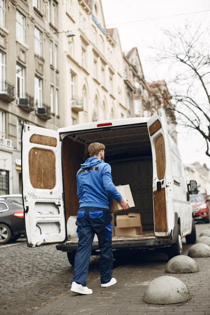 A man dressed in a dark quilted vest over a blue long-sleeved top, wearing a black and grey cap, stands outside a white van with an open rear door. He is holding a small object in his gloved hands and