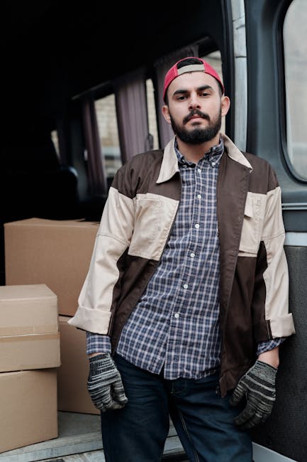 A man with a beard and wearing a red baseball cap, a beige and brown patchwork jacket over a blue checkered shirt, and grey work gloves stands inside the back of a moving van. The van's interior shows several cardboard moving boxes stacked behind him, with some boxes partially visible on the pavement outside the vehicle’s open sliding door. The background beyond the van includes a blurred street scene. This image illustrates the loading process during a home relocation, capturing a moment of furniture transport and packing for a house move, consistent with services offered by Man with Van Dulwich for quick and efficient moves in East Dulwich SE22.