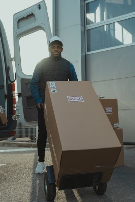 A man with dark skin, wearing a dark blue jacket, black trousers, white sneakers, and a light grey cap, stands next to a large cardboard box labeled 'Fragile' and 'Handle with Care' on a black hand truck, positioned outside a white delivery van with its side door open. In the background, there is a modern building with large glass windows and metal framing, indicating the setting is part of a home relocation or furniture transport process. The loading area appears well-lit with natural daylight. The presence of packed boxes and the loading activity suggests a professional moving or removals service, consistent with the offerings of Man with Van Dulwich, supporting efficient packing and moving logistics.