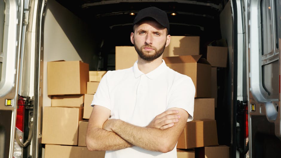A man with a beard and wearing a red baseball cap, a beige and brown patchwork jacket over a blue checkered shirt, and grey work gloves stands inside the back of a moving van. The van's interior shows several cardboard moving boxes stacked behind him, with some boxes partially visible on the pavement outside the vehicle’s open sliding door. The background beyond the van includes a blurred street scene. This image illustrates the loading process during a home relocation, capturing a moment of furniture transport and packing for a house move, consistent with services offered by Man with Van Dulwich for quick and efficient moves in East Dulwich SE22.